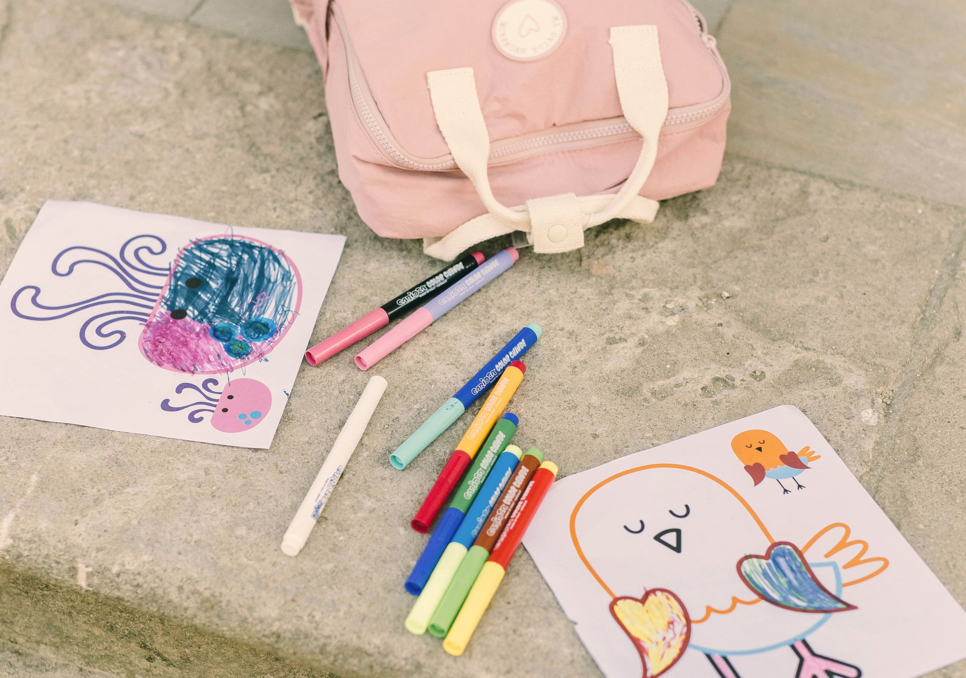 Värikyniä, piirrettyjä värityskuvia mustekalasta ja linnusta sekä vaaleanpunainen reppu harmaalla kivipinnalla.