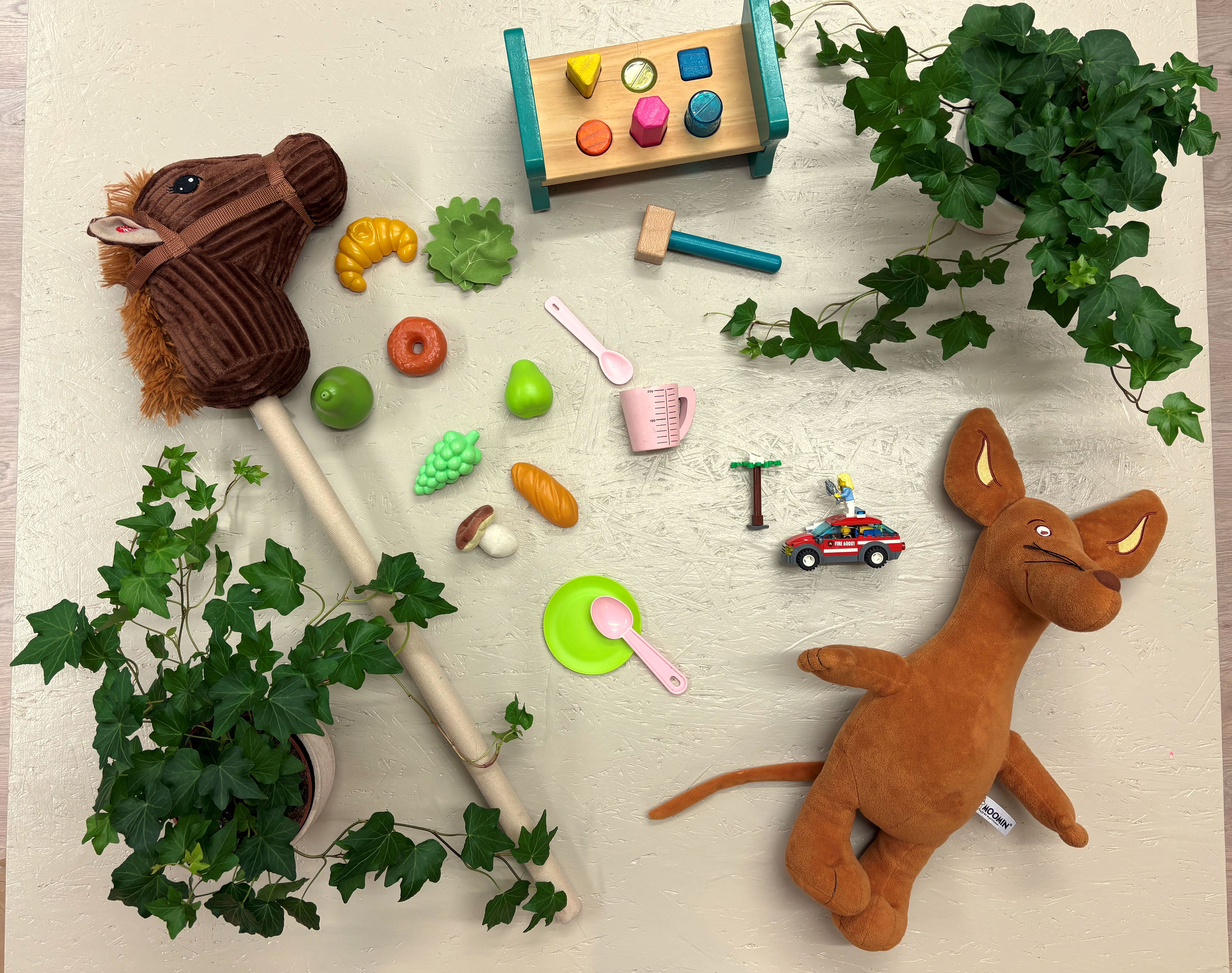 Lasten puisia leluja, pehmoleluja ja vihreitä kasveja vaalealla taustalla.